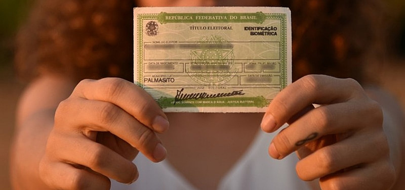 Mulher segurando título de eleitor, documento essencial para votar nas eleições. O rosto dela não aparece, só o documento, em primeiro plano. Crédito: Divulgação / TRE