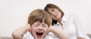 Menino gritando de olhos fechados, mãos tapando os ouvidos, em claro comportamento de birra ou pirraça no "terrible two". Mãe observando, ao fundo.