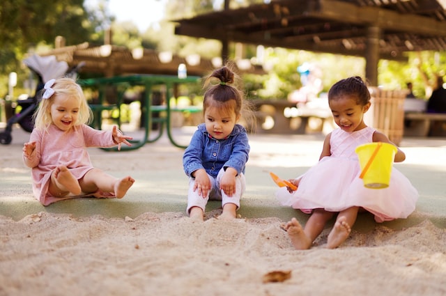 Três crianças pequenas brincando na areia, uma delas com balde na mão.