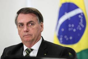 Jair Bolsonaro com a cara séria, olhando para o lado, bandeira do Brasil ao fundo, desfocada.