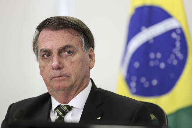 Jair Bolsonaro com a cara séria, olhando para o lado, bandeira do Brasil ao fundo, desfocada.