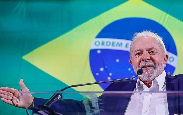 Foto de Luiz Inácio Lula da Silva, o Lula (PT), sorridente, falando, com braços abertos, e a bandeira do Brasil ao fundo.