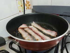 Tiras de bacon fritando em frigideira.