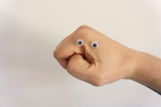 Mão fechada com dois olhos pequenos grudados nela, formando a imagem de um rosto emburrado.