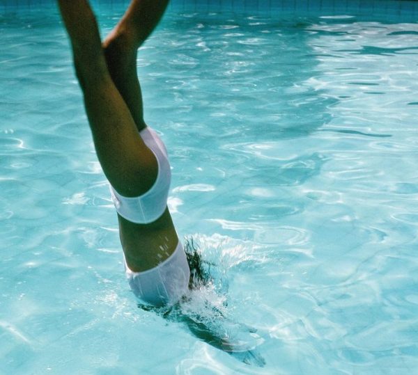 Mulher de biquíni pulando em uma piscina. Imagem em uma das capas internacionais do livro "Nadando de Volta Para Casa", de Deborah Levy.