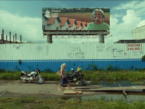 Cena do filme O Último Azul. Embaixo do cartaz com a propaganda do governo, uma pichação pede: 'Devolvam o meu avô!'